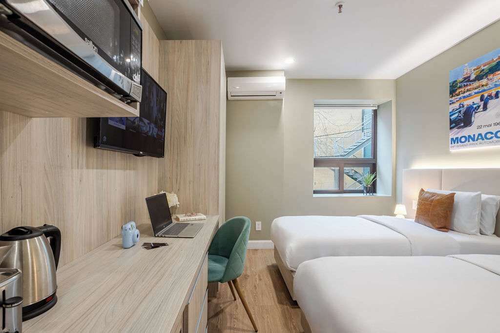 Quarto de hotel com escrivaninha de madeira, laptop, chaleira, televisão, cama e um cartaz na parede.