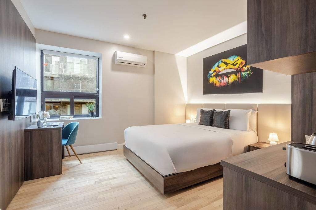 Une chambre d’hôtel moderne, équipée d’un lit, d’un bureau et d’une fenêtre donnant sur un paysage urbain.