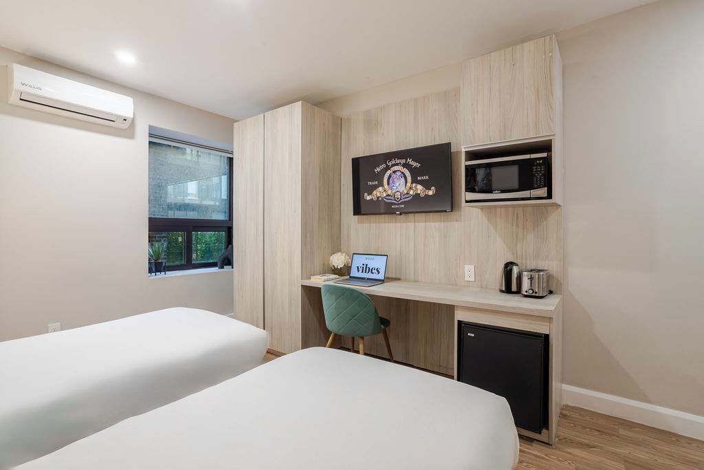 Habitación de hotel con dos camas, un escritorio con una computadora portátil, un microondas, un refrigerador pequeño y un televisor montado en la pared.