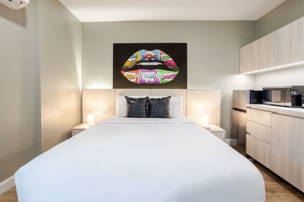 Una habitació d'hotel moderna amb un llit gran, taules de nit amb làmpades, microones i zona de cuina.
