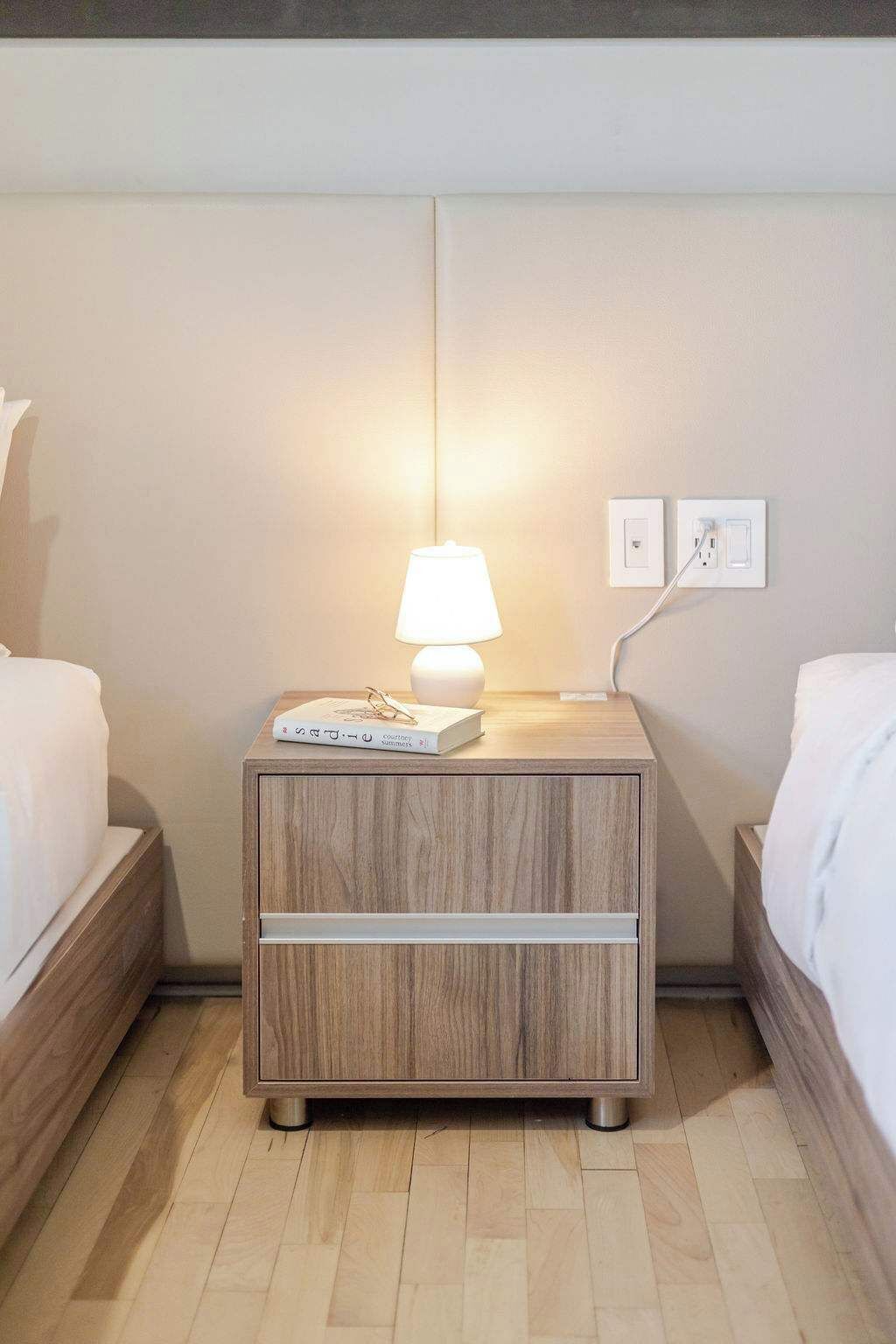 A neatly arranged hotel room with a wooden nightstand, a bedside lamp, a book, and a power outlet.