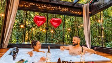 Ein Paar entspannt sich in einer Badewanne mit Blick auf viel Grün, umgeben von herzförmigen Dekorationen und Lichterketten.