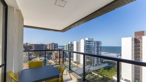 Un balcon avec une table et des chaises, donnant sur un paysage urbain côtier, avec des bâtiments modernes et un ciel dégagé.