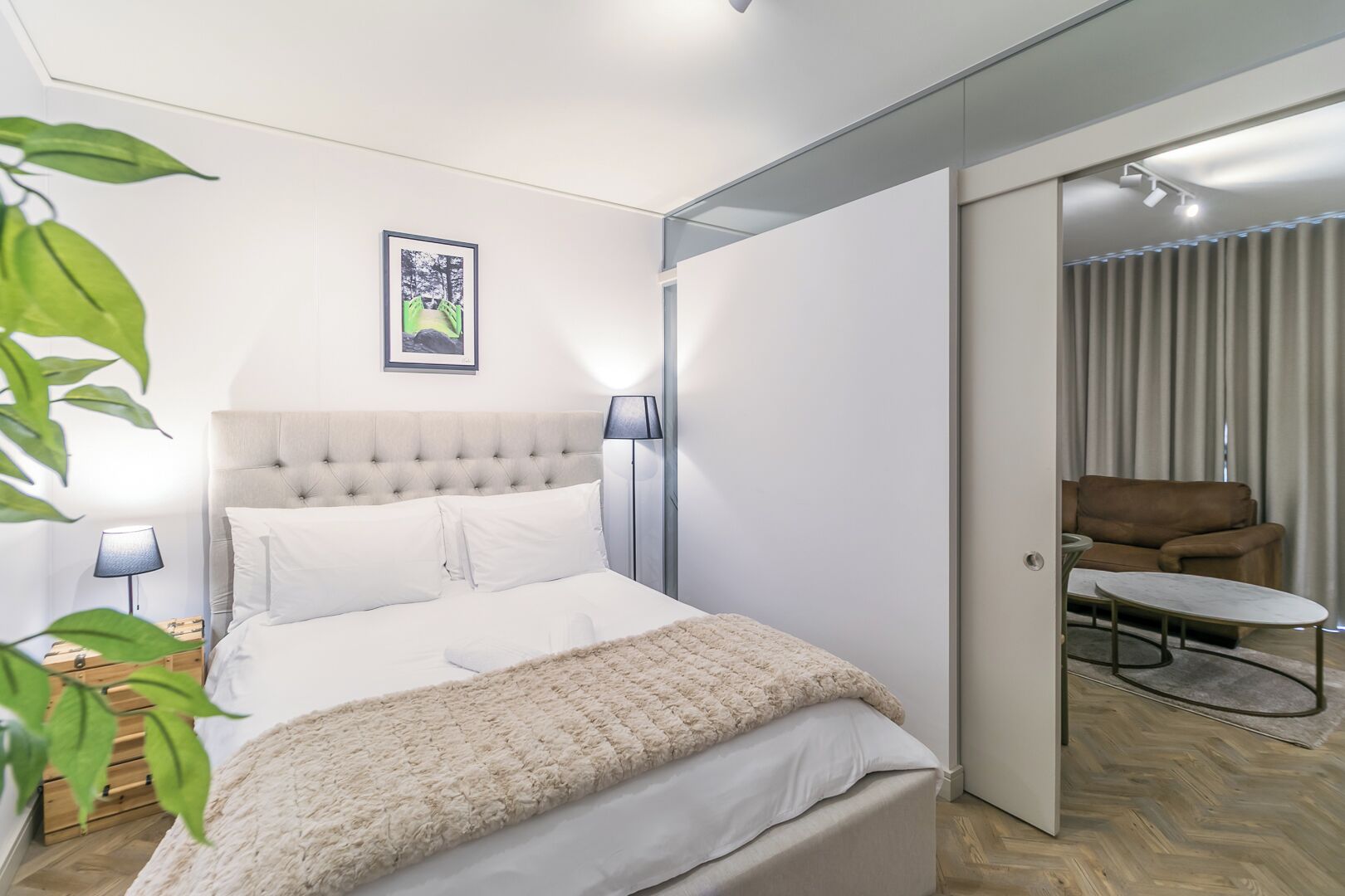 Ein modernes Schlafzimmer mit einer gepolsterten Kopfstütze, einem Bett mit weißen Bettwäsche, einer flauschigen Decke und einem Nachttisch mit einer Lampe. Ein gerahmtes Bild hängt über dem Bett, und eine Tür führt zu einem Wohnbereich mit einer Couch und einem kleinen Tisch.