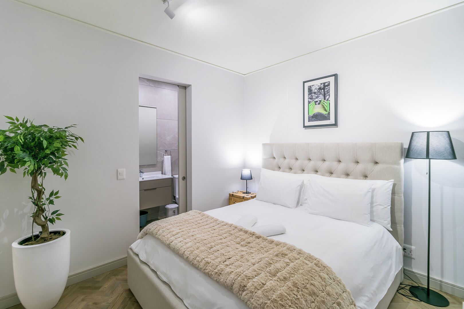 A bedroom with a bed, a bedside lamp, a nightstand, a potted plant, and a framed picture on the wall.