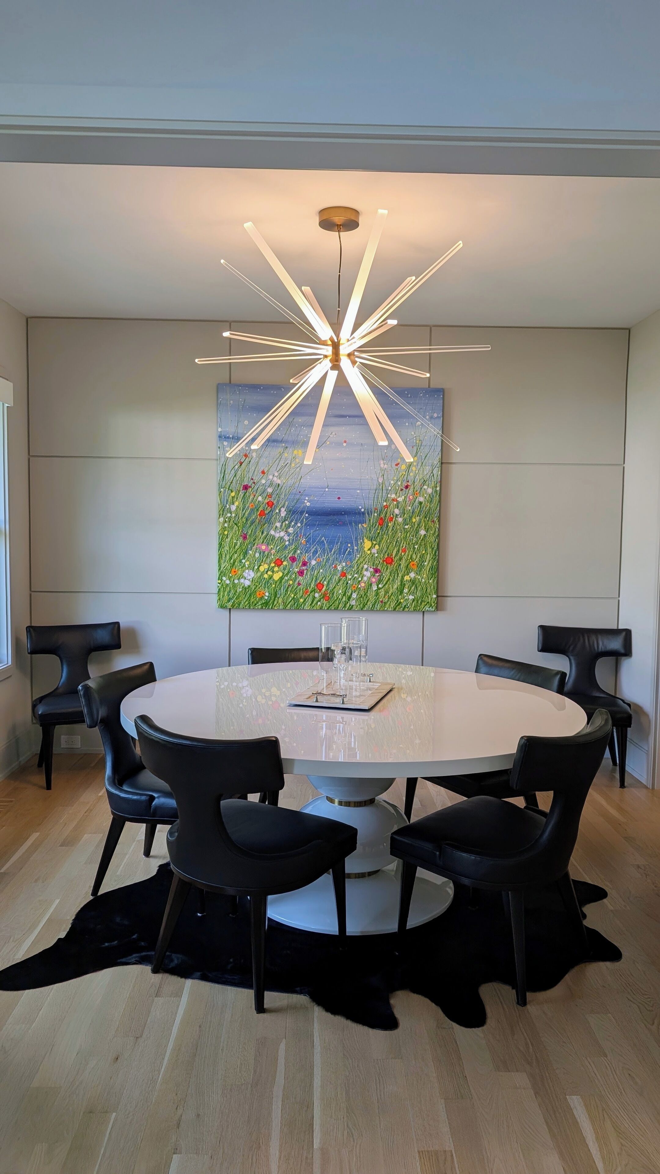 Un espace repas moderne avec une table ronde, des chaises noires, un luminaire au plafond original et un grand tableau floral accroché au mur.