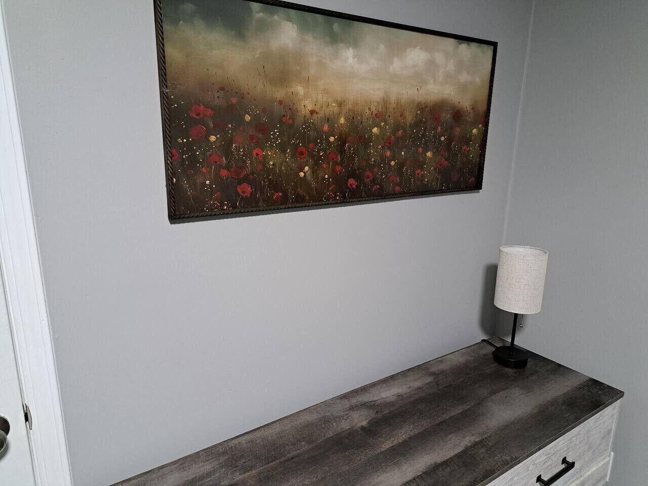 A room with a wooden bench, a painting of a field with flowers, and a lamp on the bench.