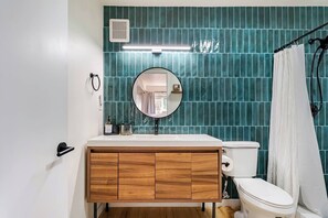 Une salle de bain avec des toilettes blanches, un meuble-lavabo en bois, un miroir rond et des murs recouverts de carreaux de couleur turquoise.