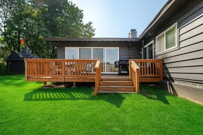 Un jardin avec une terrasse en bois, un barbecue et une maison dotée de grandes fenêtres.