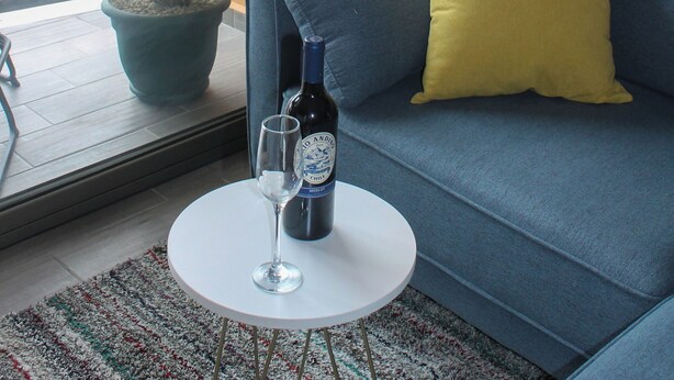 Una sala de estar moderna con un sofá azul, un cojín amarillo, una planta en maceta, una botella de vino y un vaso sobre una mesita redonda.