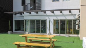 Una mesa de picnic de madera sobre césped artificial, frente a un edificio moderno con grandes ventanales y un balcón.