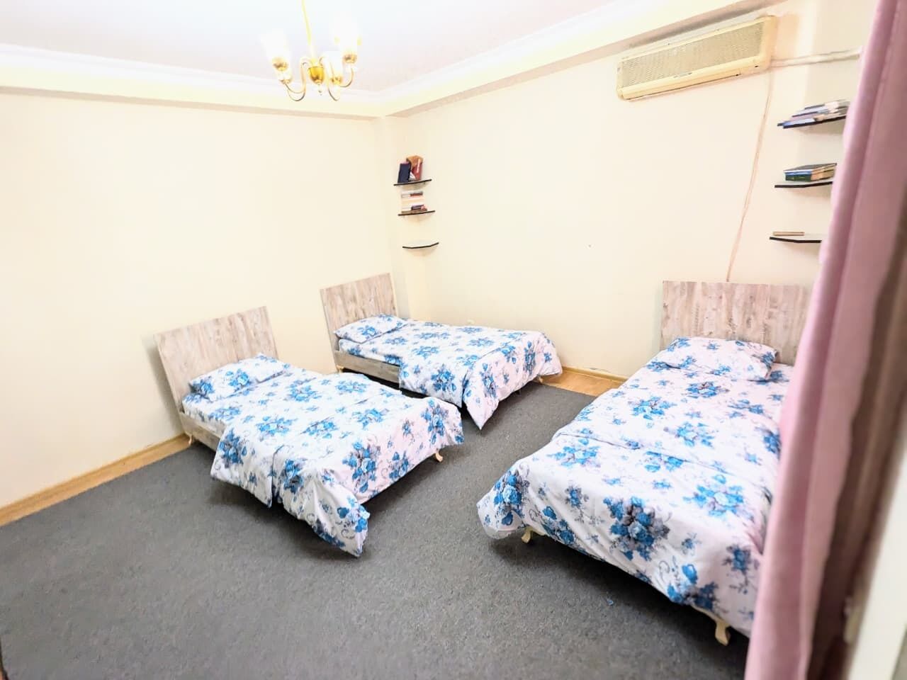 Une chambre avec trois lits simples, une tête de lit en bois, une petite étagère avec des livres et un ventilateur de plafond.