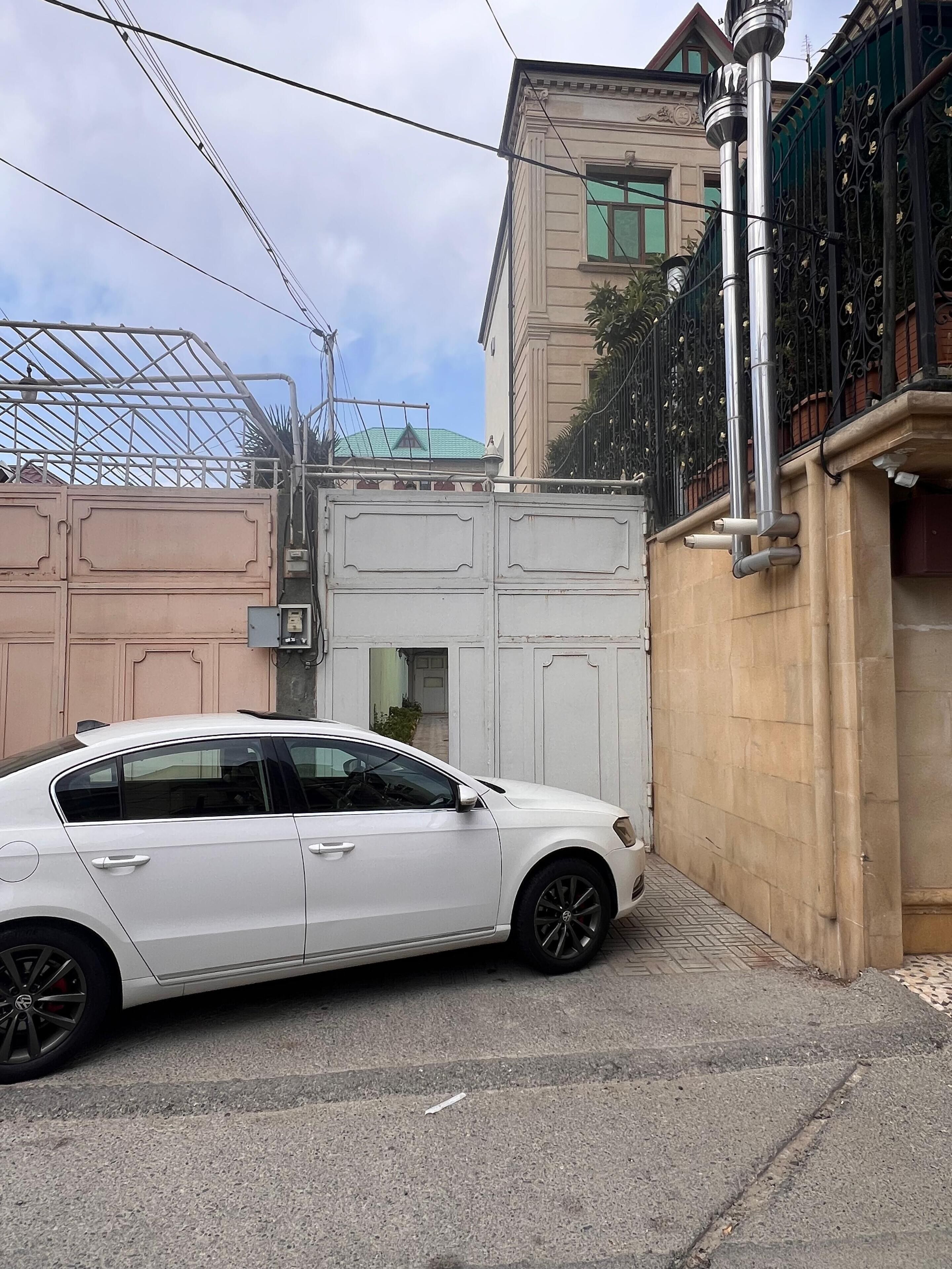 A white car parked in a narrow street with a building featuring a green roof and ornate ironwork in the background.