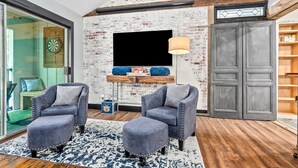 Una sala de estar moderna con pared de ladrillo, un televisor de pantalla plana, dos sillones grises, un tapete azul y blanco y piso de madera.