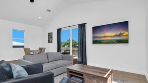 A modern living room with a grey sofa, a black leather couch, and a wooden coffee table. There is a dining area with a table and chairs, and a large window with curtains. - Harbor Place Boathouse, BRAND NEW DOCK, Open, on stilts, ocean front (Marco Island)
