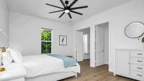 A bedroom with a bed, bedside table, dresser, and a ceiling fan. There is a window with a view of trees and a door leading to another room. - Harbor Place Boathouse, BRAND NEW DOCK, Open, on stilts, ocean front (Marco Island)