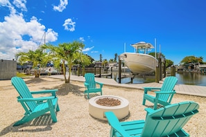 A waterfront area with a boat docked, surrounded by turquoise Adirondack chairs, a fire pit, and palm trees. - Harbor Place Boathouse, BRAND NEW DOCK, Open, on stilts, ocean front (Marco Island)