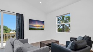 A modern living room with a grey sofa, a black armchair, and a wooden coffee table. There is a painting of a sunset on the wall and a window with a view of palm trees. - Harbor Place Boathouse, BRAND NEW DOCK, Open, on stilts, ocean front (Marco Island)