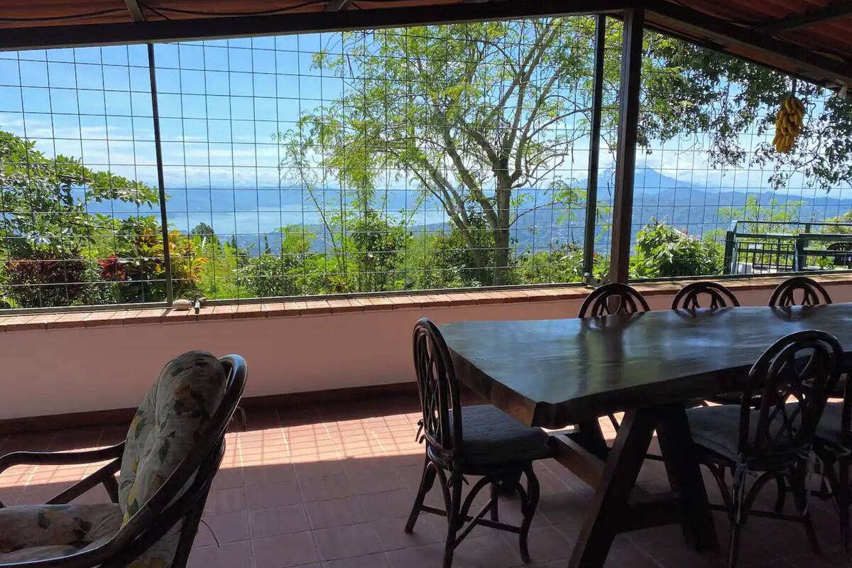 A covered outdoor seating area with a table and chairs, overlooking a scenic view of mountains and greenery.