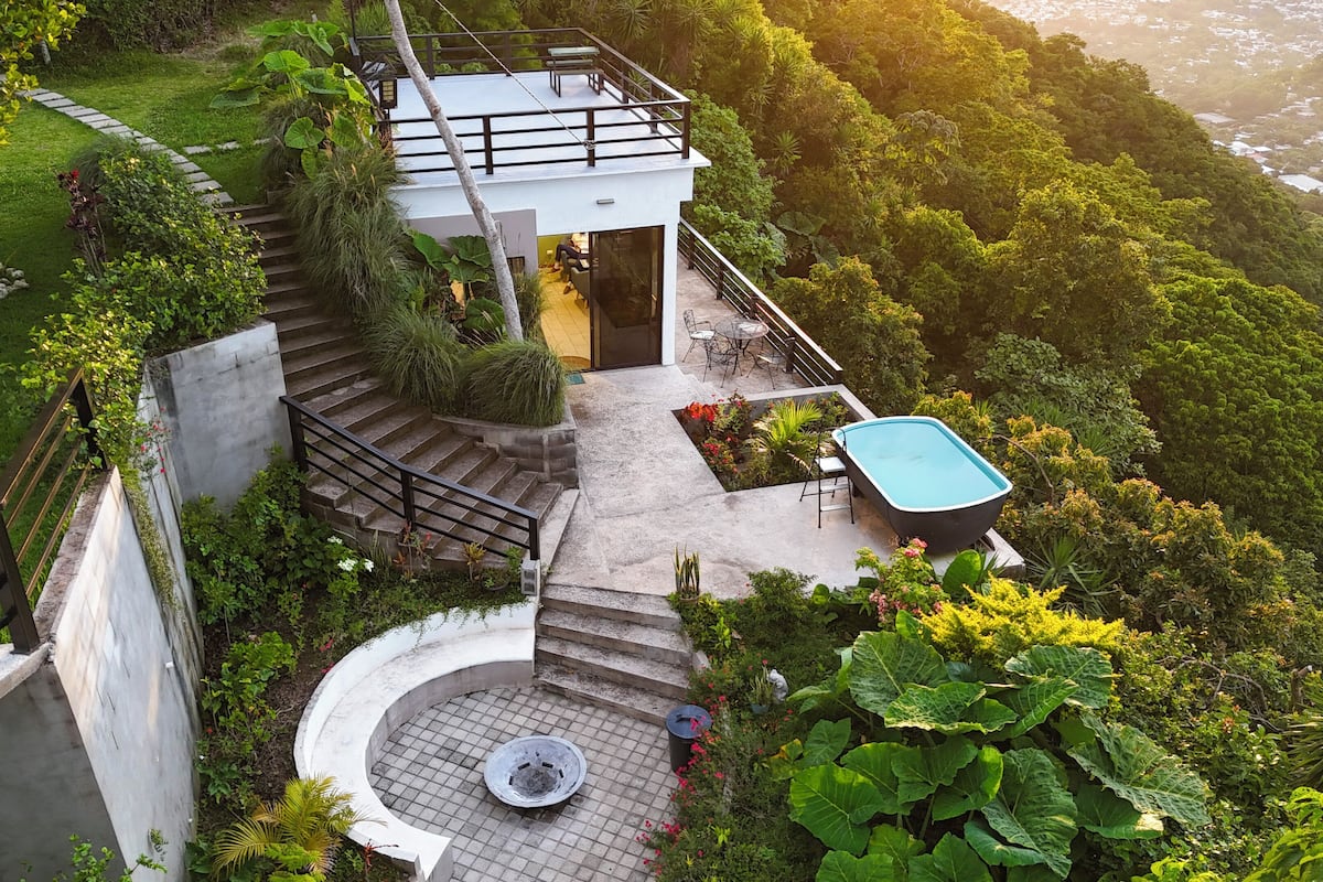 A modern house with a circular staircase, a pool, and lush greenery surrounding it.