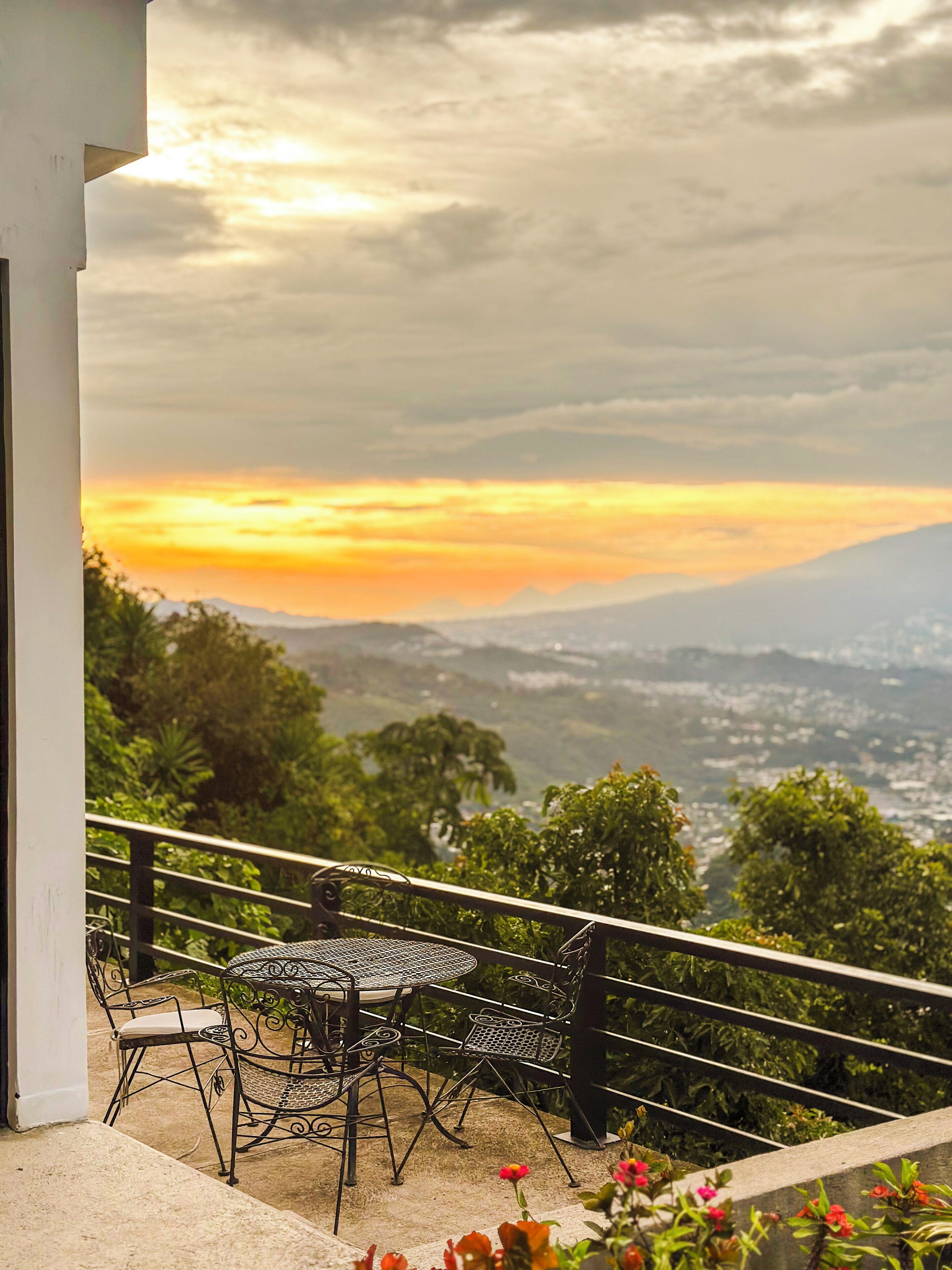 Un balcon avec une table et des chaises, offrant une vue panoramique sur un paysage magnifique, avec des montagnes et un coucher de soleil.