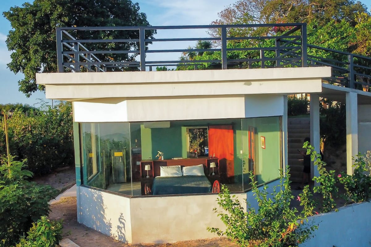 A modern glass-walled bedroom with a bed, a desk, and a staircase leading to an upper level.