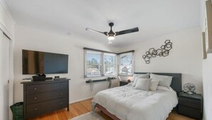 Een slaapkamer met een bed, een dressoir, een televisie, een plafondventilator en een raam met jaloezieën.