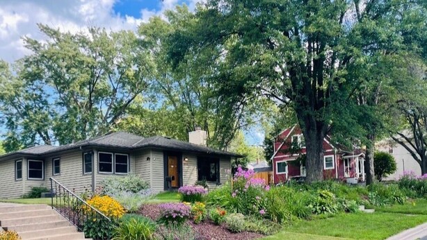 Een tweelaags huis met een veranda, een kleine voortuin vol planten, en een oprit naar de voordeur.