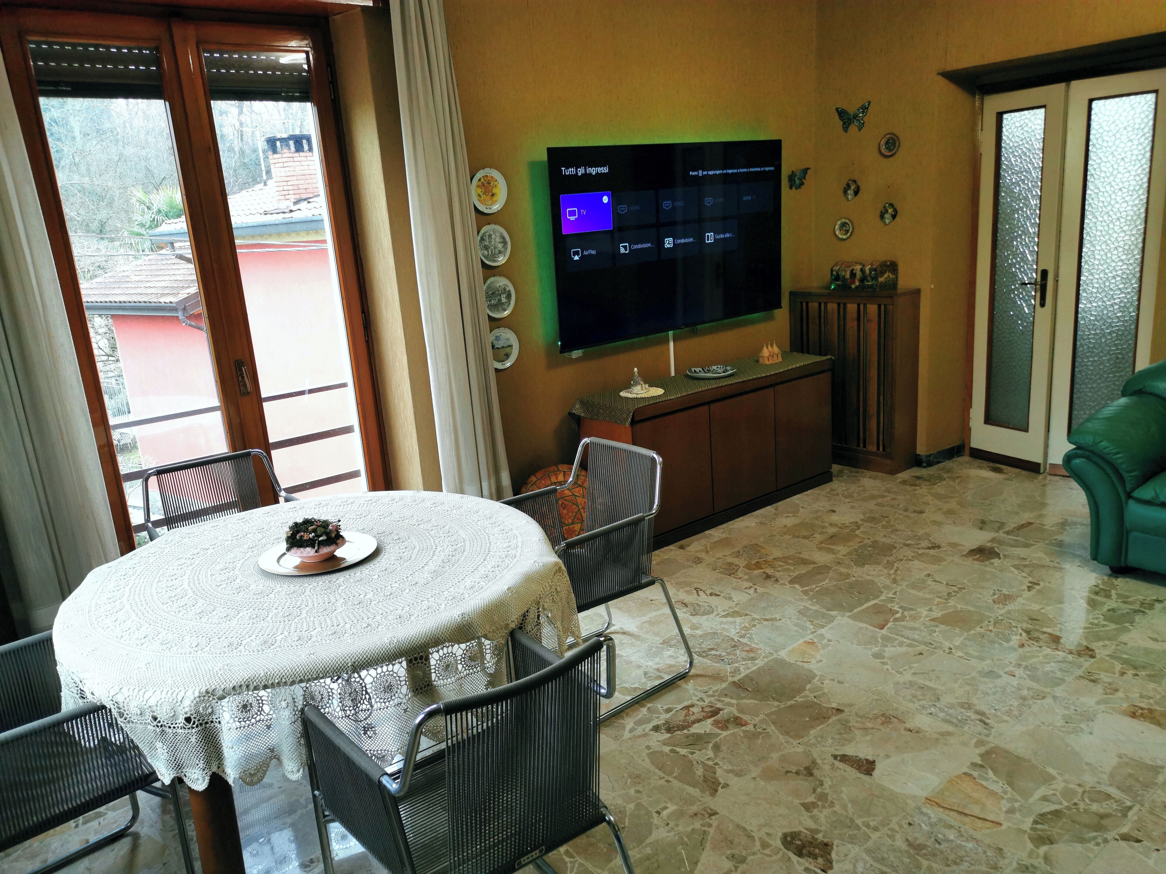 A dining area with a round table covered by a white lace tablecloth, a black metal chair, and a small decorative bowl. A television is mounted on the wall, and there are decorative plates and a small cabinet below it.