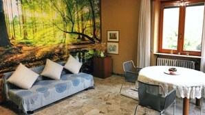 A room with a forest mural, a sofa with cushions, a dining table with chairs, and a window with curtains. - Large apartment near the lake and beach with mountain views in the greenery (Lombardia)