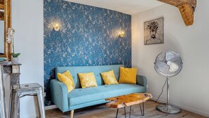A room with a blue textured wall, a teal sofa with yellow cushions, a wooden coffee table, a standing fan, and a framed artwork on the wall.