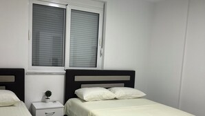 A bedroom with two beds, a nightstand, a window with blinds, and a ceiling light. - A family owned house (Ulcinj)