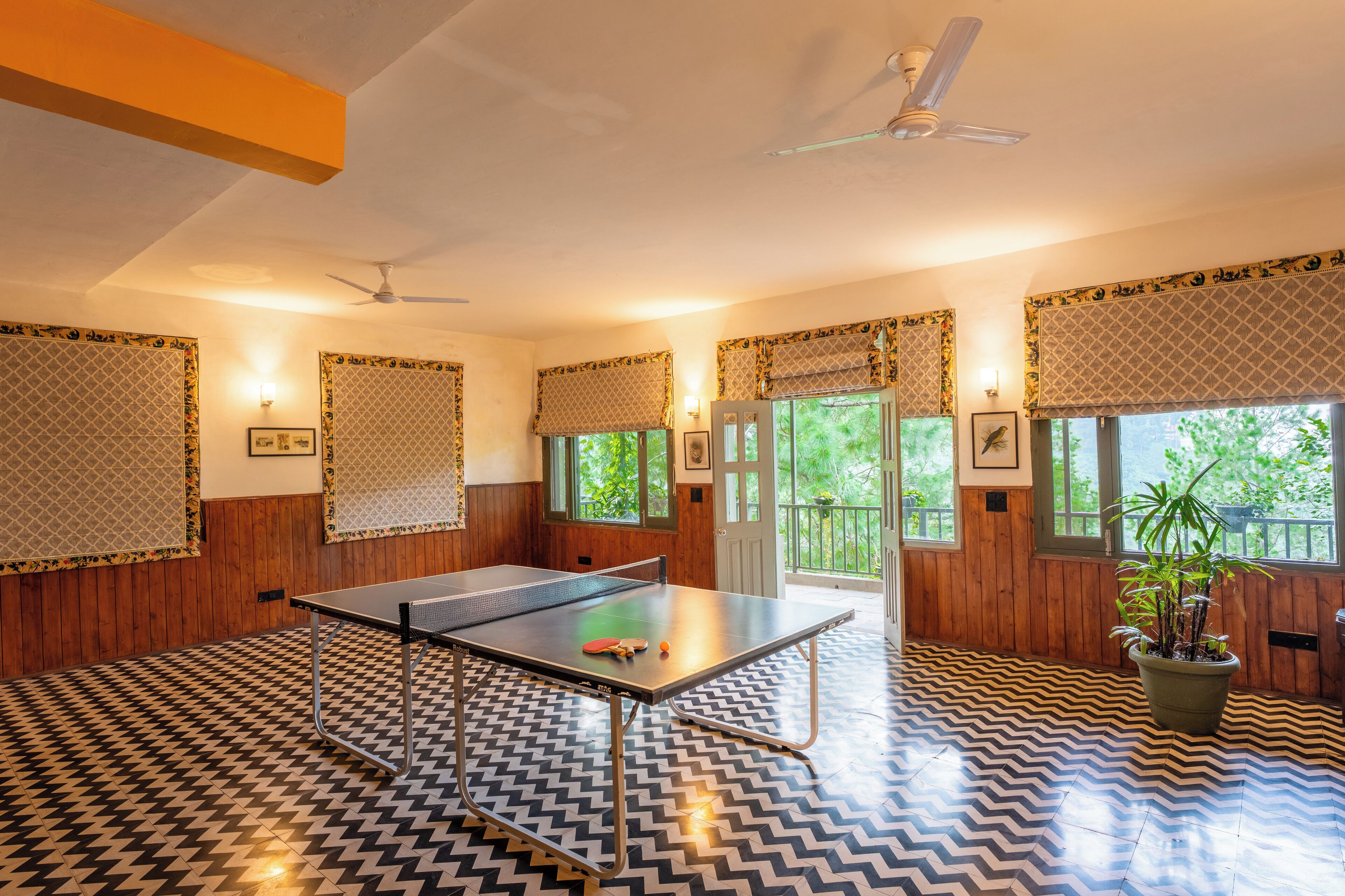 Um quarto com mesa de pingue-pongue, paredes de madeira, piso estampado, ventiladores de teto e janelas com persianas.