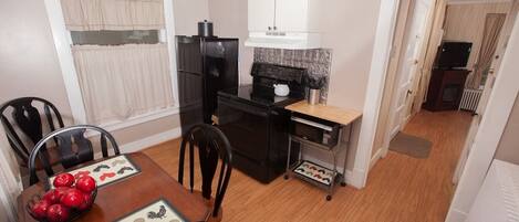 A compact kitchen with a refrigerator, stove, and microwave. There is a dining table with chairs and a small oven cart.