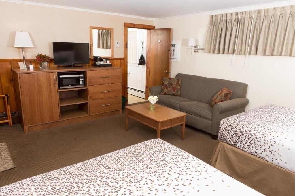 A hotel room with a wooden desk, a television, a sofa, two beds with patterned bedding, a small table with a vase, and a mirror on the wall.