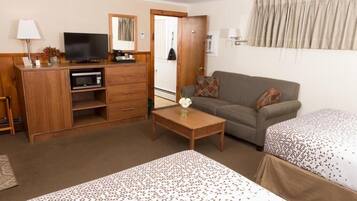 A hotel room with a wooden desk, a television, a sofa, two beds with patterned bedding, a small table with a vase, and a mirror on the wall.