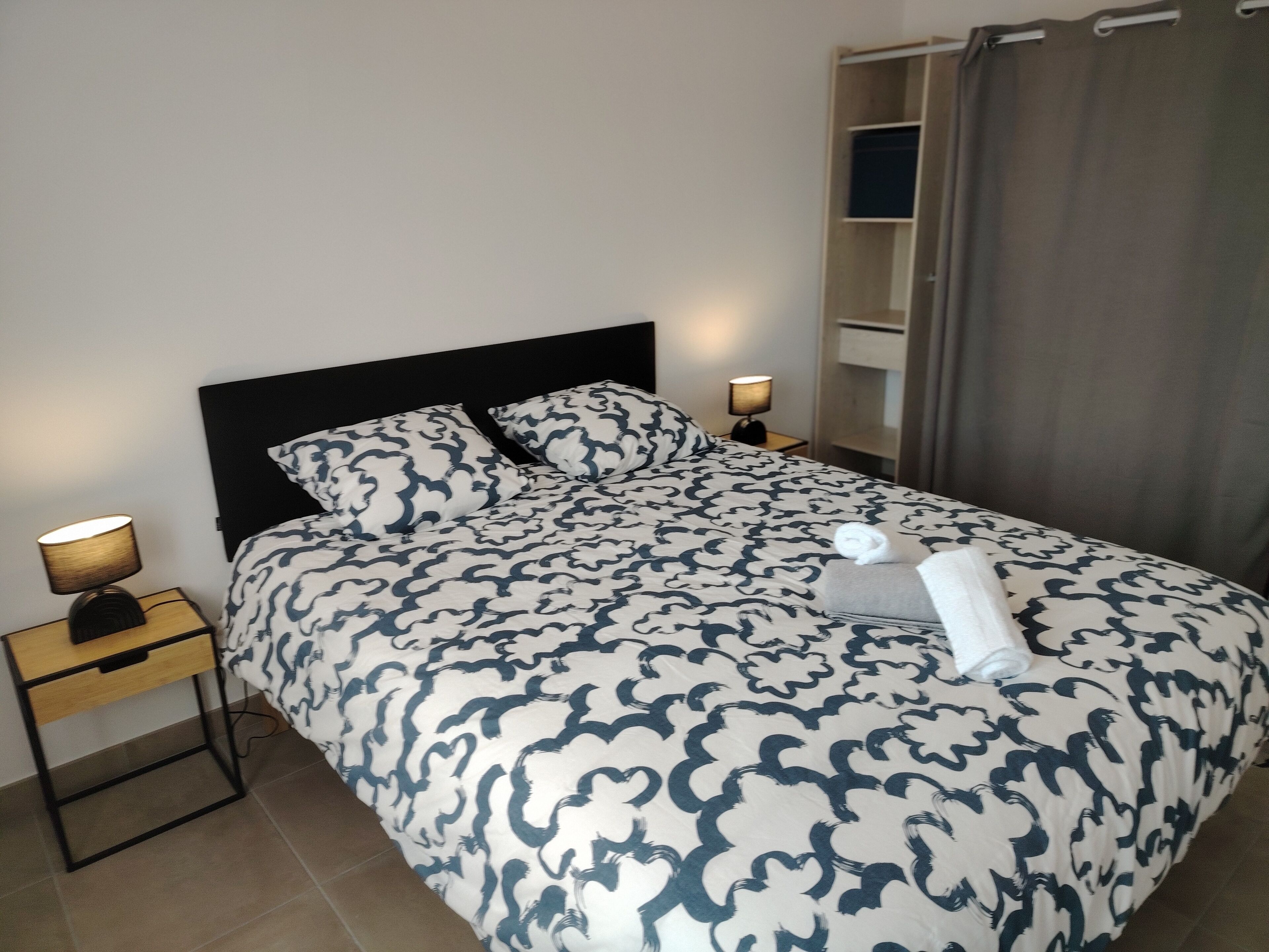 A neatly made bed with a patterned bedspread, two bedside lamps, a small wooden nightstand, and a grey curtain partially drawn.