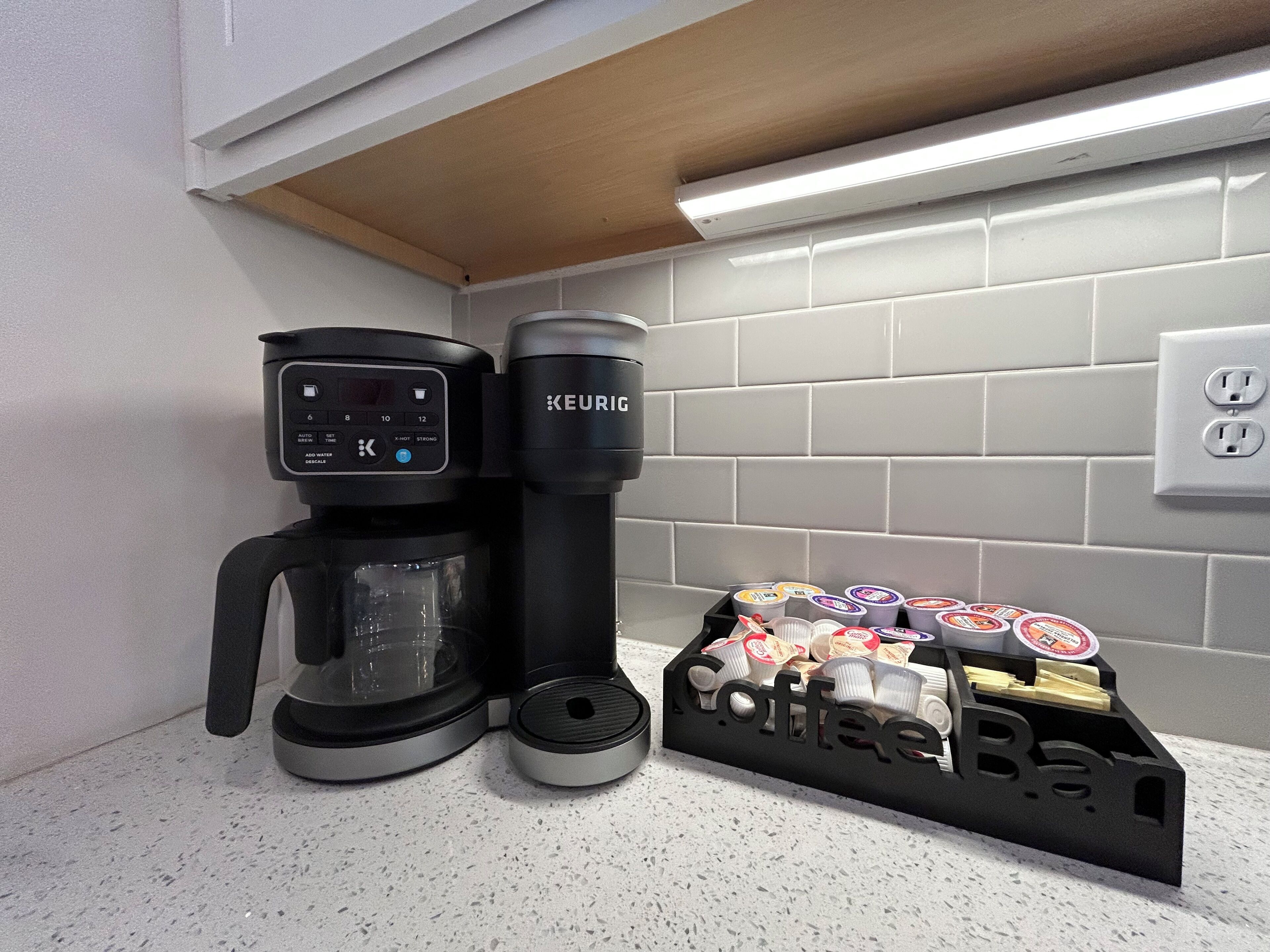Uma cafeteira, um dispensador de cápsulas de café e uma caixa de cápsulas em uma bancada de cozinha.