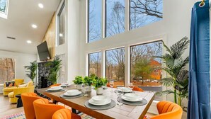 Un espace repas moderne avec une longue table en bois, dressée pour quatre personnes, entourée de chaises orange, de grandes fenêtres et de plantes d’intérieur.