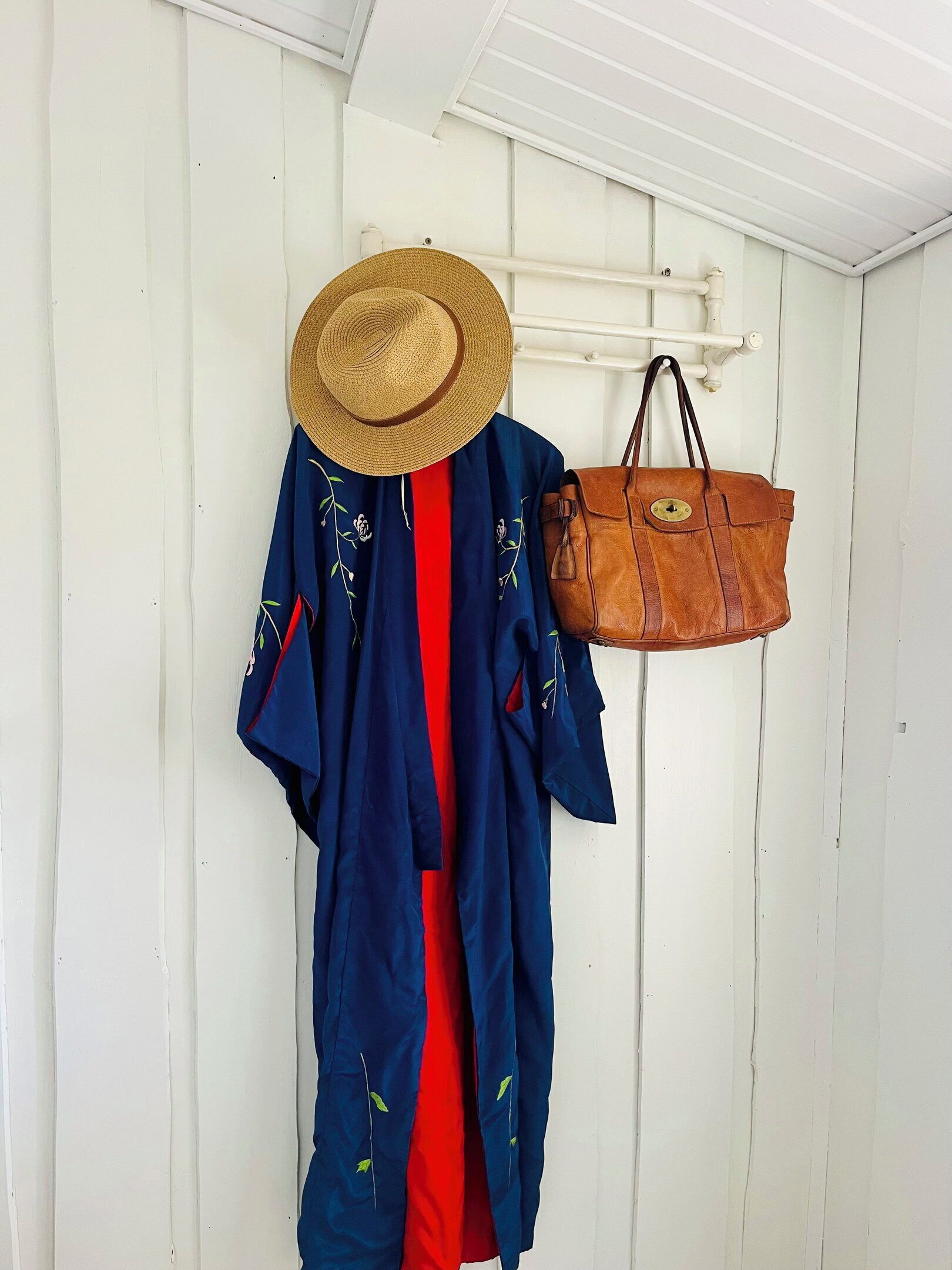 Una chaqueta azul con bordados florales, un sombrero de paja y un bolso de cuero marrón colgados en una pared blanca.