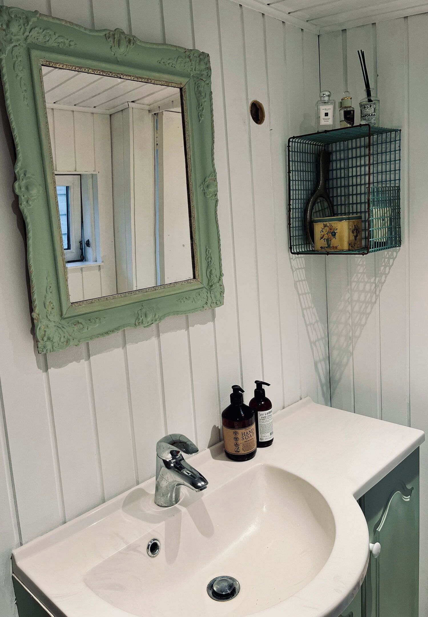 Une salle de bain avec un lavabo blanc, un miroir encadré de vert et une petite étagère contenant des articles de toilette.