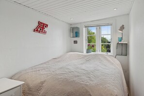 A bedroom with a bed, a window with a view, a wall-mounted lamp, and a framed picture.