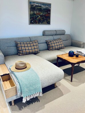 A living room with a grey sofa, plaid cushions, a woven hat, a blue throw, a wooden coffee table, and a painting on the wall.