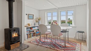 A cozy room with a wood-burning stove, a dining table with chairs, a bookshelf, and a window with a view.