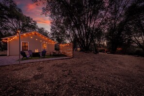 Pequeña casa con fogón y asientos exteriores, rodeada de árboles y una valla de madera.