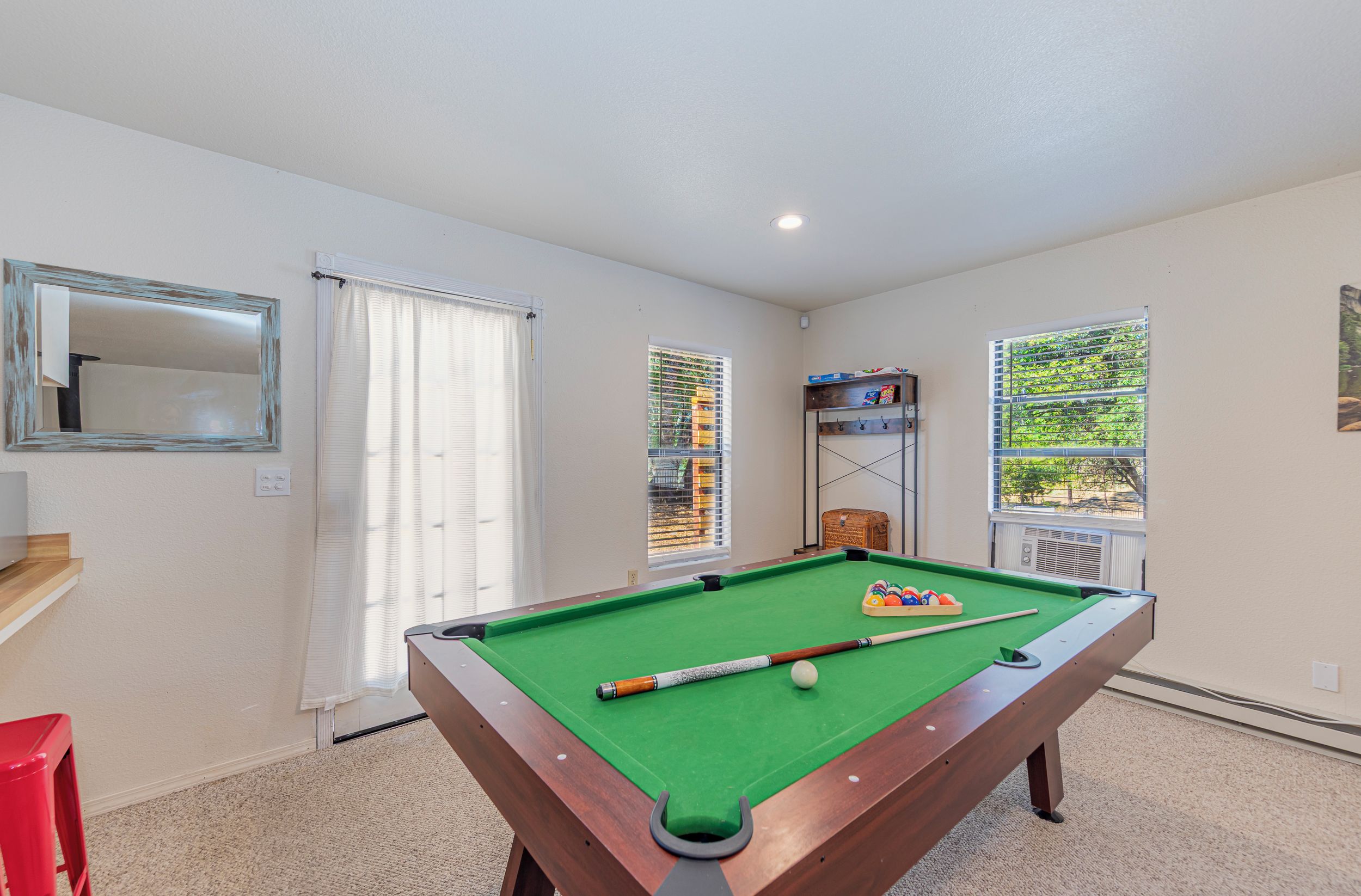 A pool table with balls and cues, a red chair, a wooden bench, and a window with blinds.