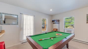 A pool table with balls and cues, a red chair, a wooden bench, and a window with blinds.