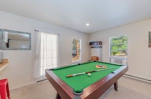 A pool table with balls and cues, a red chair, a wooden bench, and a window with blinds.