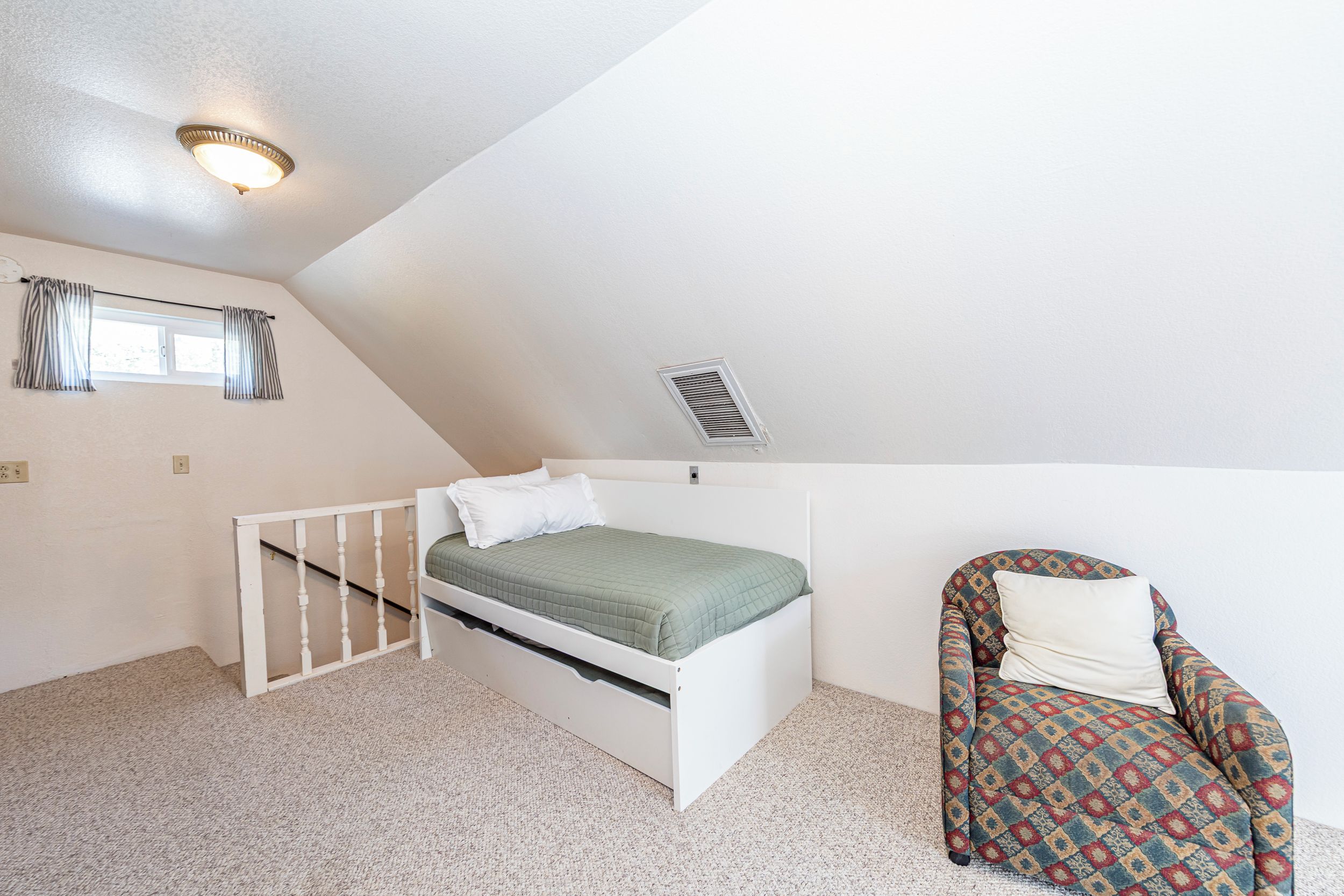 A small, single-bed room with a sloped ceiling, a window with curtains, a radiator, and a patterned armchair.