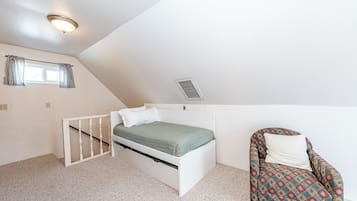 A small, single-bed room with a sloped ceiling, a window with curtains, a radiator, and a patterned armchair.
